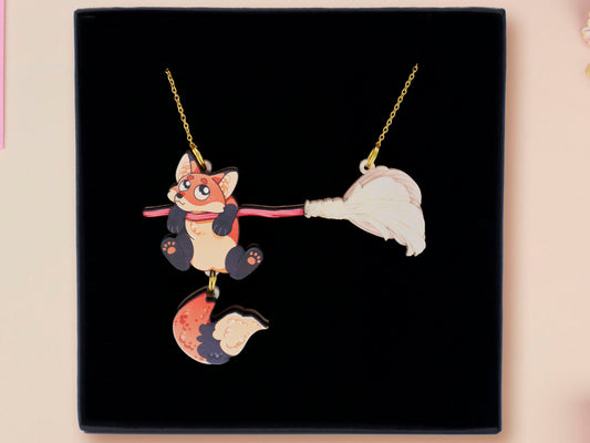 Mixed material handmade necklace of chibi cartoon Fox hanging on to a pearlescent witches broomstick, with a gold chain and black gift box.