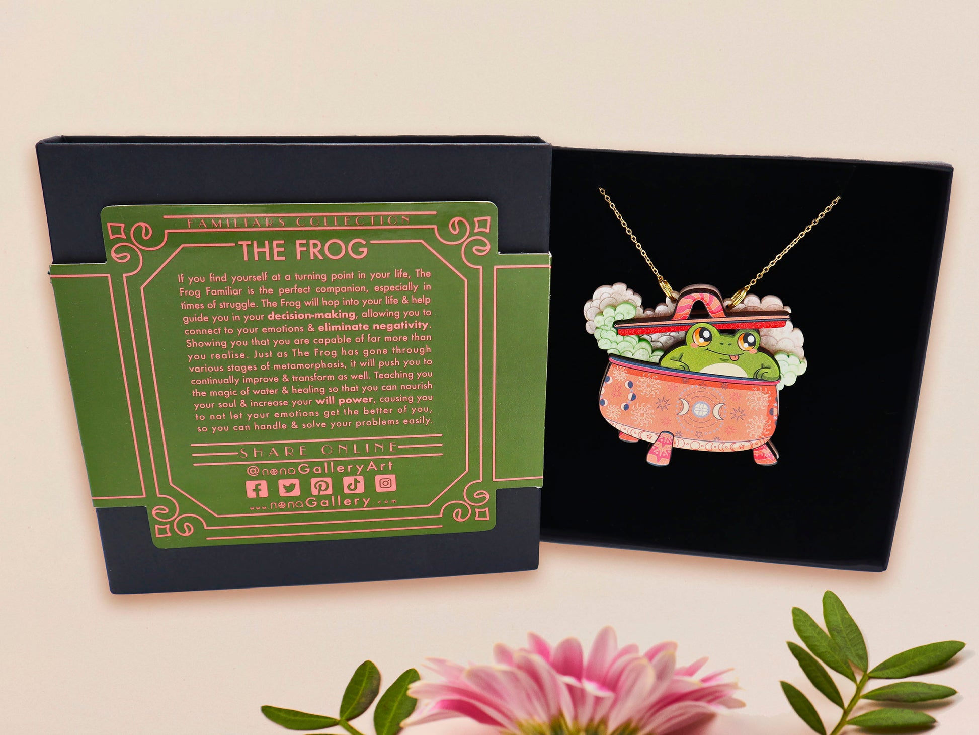 Mixed material handmade necklace of chibi cartoon frog sat inside a bubbling pearlescent witch's cauldron, with a gold chain and black gift box with a green familiars collection gift sleeve.