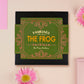 A wooden and glitter acrylic necklace with gold chain in a black box of two cute frogs sat on a log smiling surrounded by flowers, plants and mushrooms