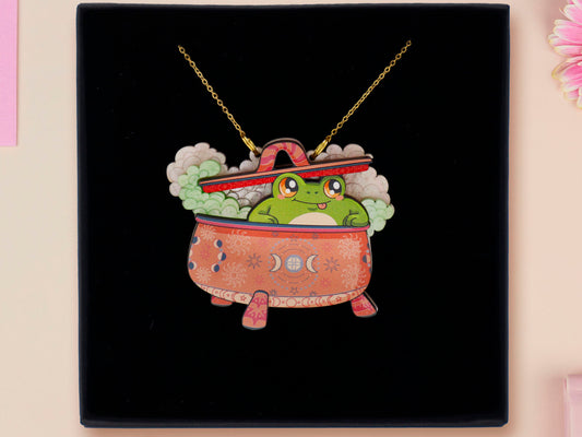 Mixed material handmade necklace of chibi cartoon frog sat inside a bubbling pearlescent witch's cauldron, with a gold chain and black gift box.