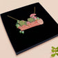 A wooden and glitter acrylic necklace with gold chain in a black box of two cute frogs sat on a log smiling surrounded by flowers, plants and mushrooms
