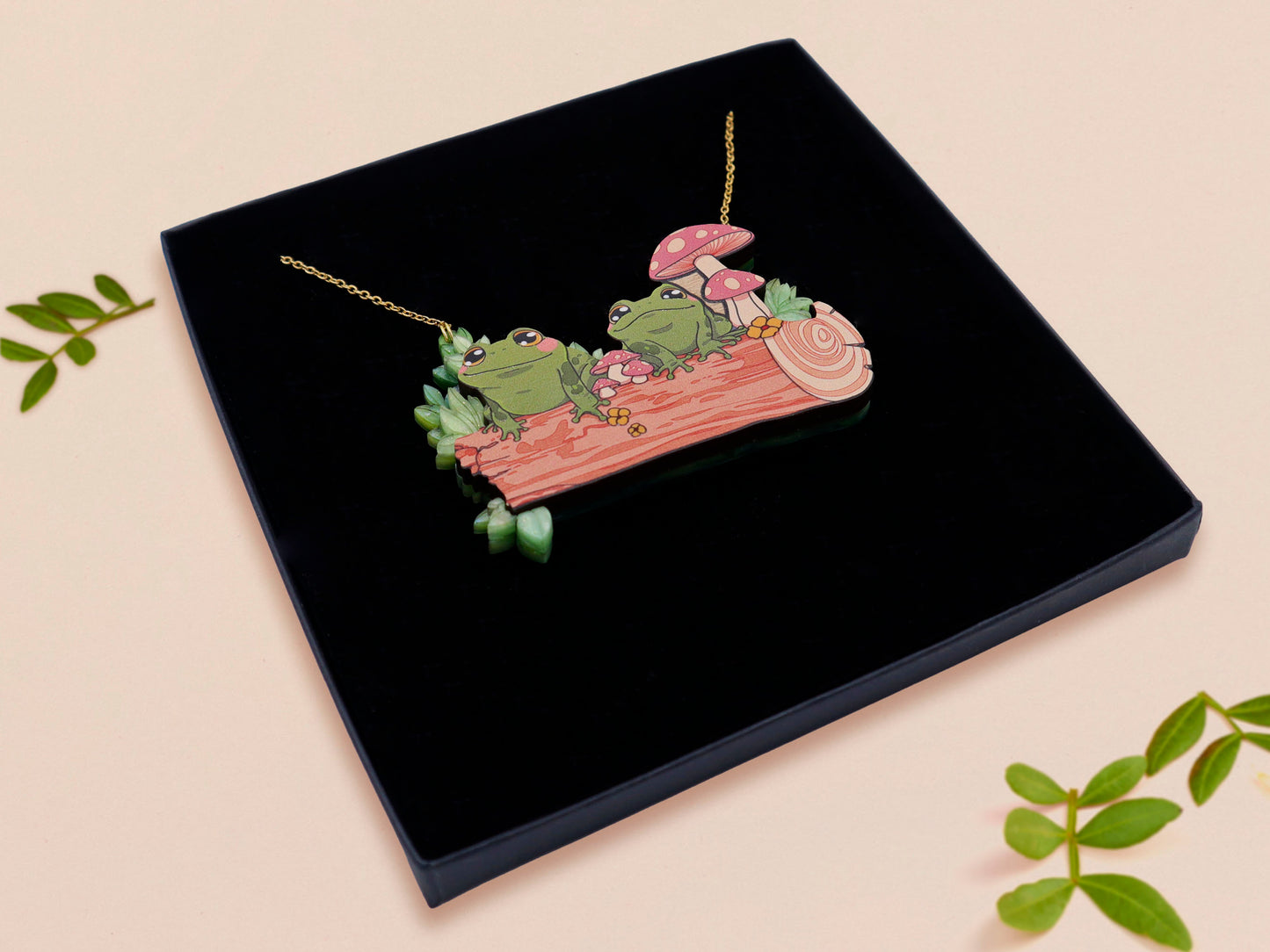 A wooden and glitter acrylic necklace with gold chain in a black box of two cute frogs sat on a log smiling surrounded by flowers, plants and mushrooms