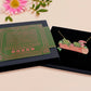 A wooden and glitter acrylic necklace with gold chain in a black box of two cute frogs sat on a log smiling surrounded by flowers, plants and mushrooms
