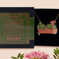 A wooden and glitter acrylic necklace with gold chain in a black box of two cute frogs sat on a log smiling surrounded by flowers, plants and mushrooms