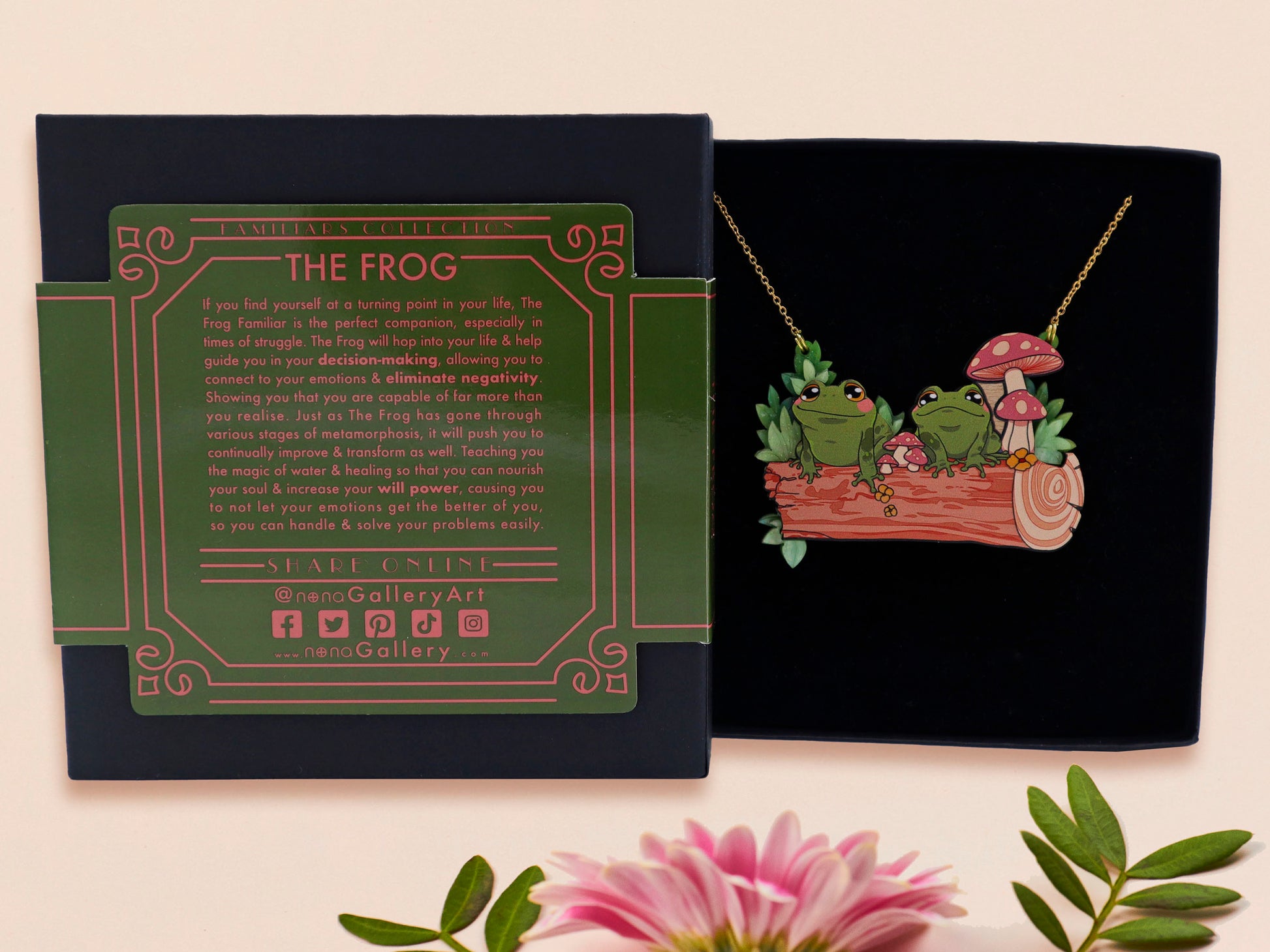 A wooden and glitter acrylic necklace with gold chain in a black box of two cute frogs sat on a log smiling surrounded by flowers, plants and mushrooms