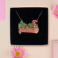 A wooden and glitter acrylic necklace with gold chain in a black box of two cute frogs sat on a log smiling surrounded by flowers, plants and mushrooms