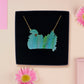 A wooden and glitter acrylic necklace with gold chain in a black box of two cute frogs sat on a log smiling surrounded by flowers, plants and mushrooms