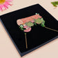 A wooden and glitter acrylic necklace with gold chain in a black box of two cute frogs sat on a log smiling surrounded by flowers, plants and mushrooms