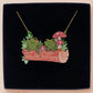 A wooden and glitter acrylic necklace with gold chain in a black box of two cute frogs sat on a log smiling surrounded by flowers, plants and mushrooms