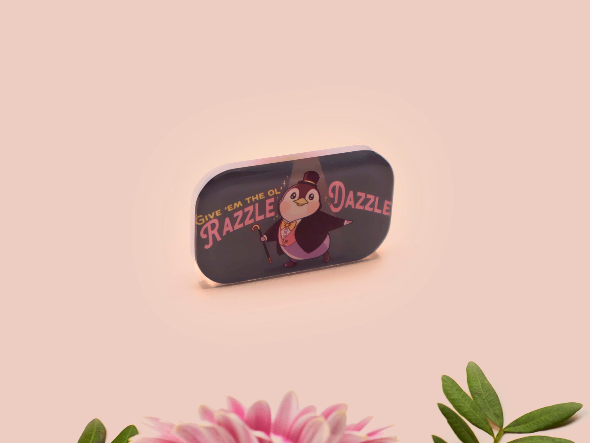 A clear acrylic pin badge with a pink heart-shaped pin back clasp. The design of the pin badge is of a cute chibi penguin dancing in a suit under a spotlight with the quote give 'em the ol' razzle dazzle