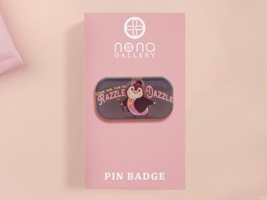 A clear acrylic pin badge with a pink heart-shaped pin back clasp. The design of the pin badge is of a cute chibi penguin dancing in a suit under a spotlight with the quote give 'em the ol' razzle dazzle