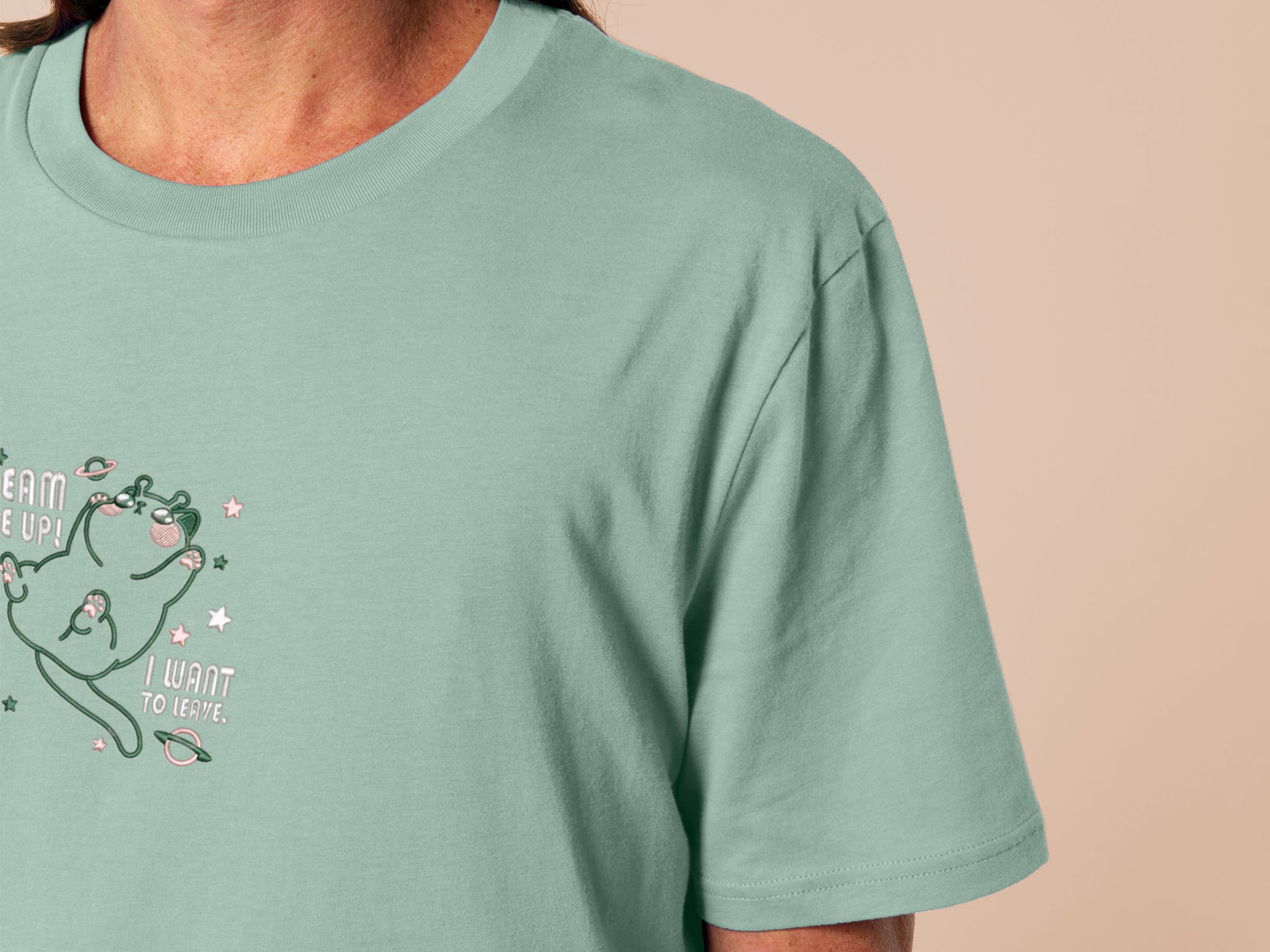 An embroidered green t-shirt design of a cute alien cat floating in space surrounded by stars and planets and by the quote beam me up! I want to leave