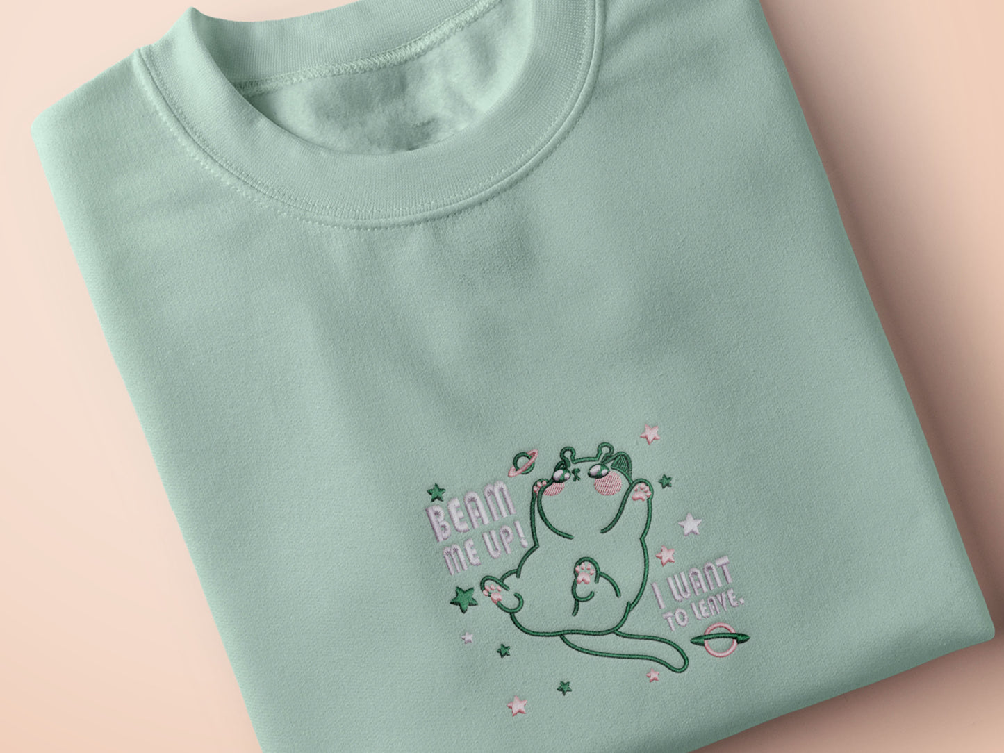 An embroidered green t-shirt design of a cute alien cat floating in space surrounded by stars and planets and by the quote beam me up! I want to leave