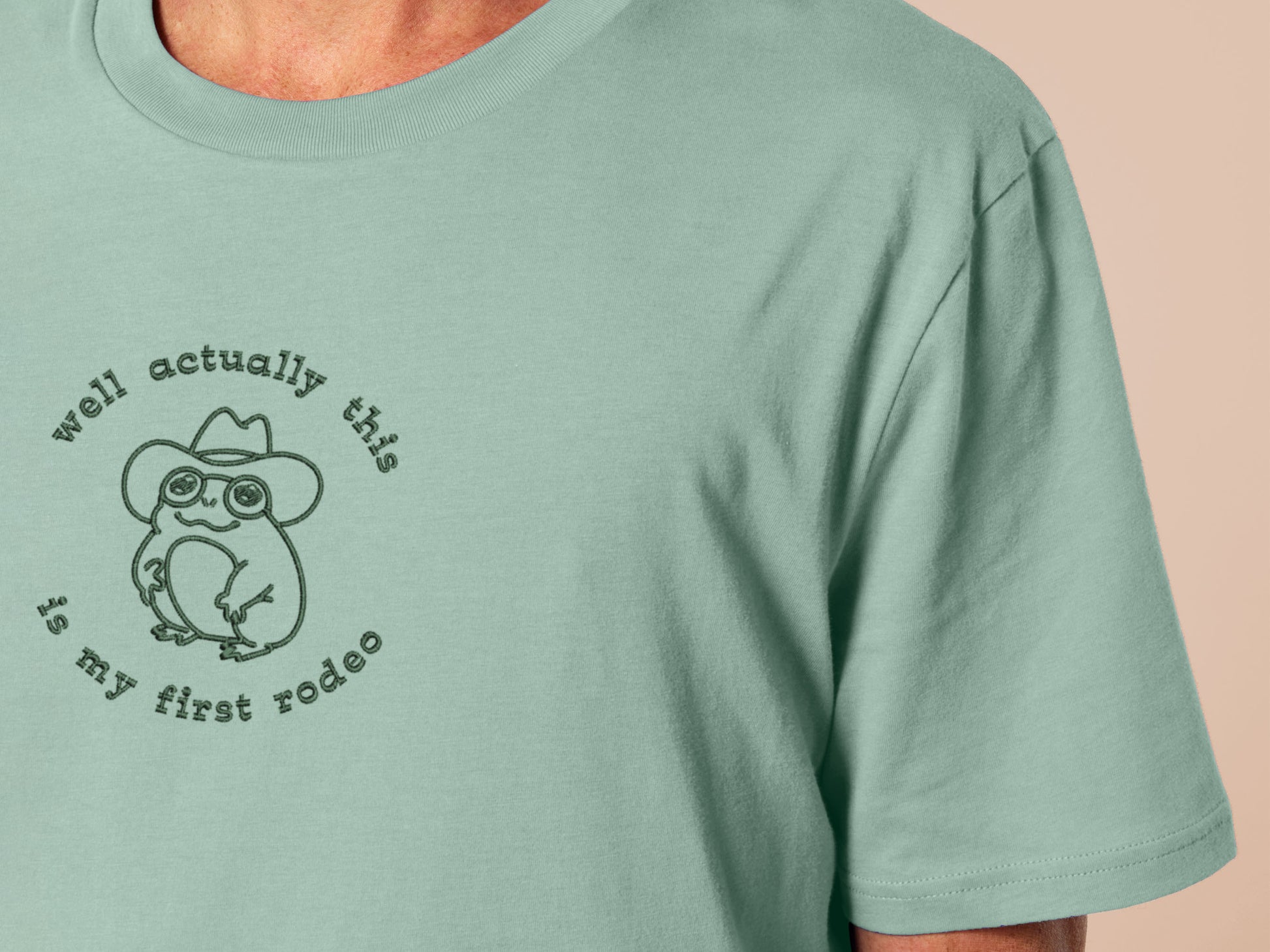 An embroidered green t-shirt design of a cute cowboy frog wearing a stetson surrounded by the test well actually this is my first rodeo.