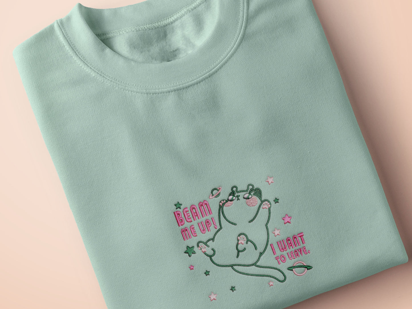 An embroidered green t-shirt design of a cute alien cat floating in space surrounded by stars and planets and by the quote beam me up! I want to leave