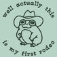 An embroidered green t-shirt design of a cute cowboy frog wearing a stetson surrounded by the test well actually this is my first rodeo.
