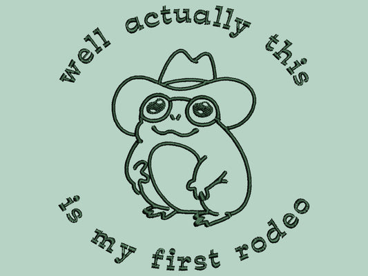 An embroidered green t-shirt design of a cute cowboy frog wearing a stetson surrounded by the test well actually this is my first rodeo.