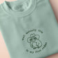 An embroidered green t-shirt design of a cute cowboy frog wearing a stetson surrounded by the test well actually this is my first rodeo.