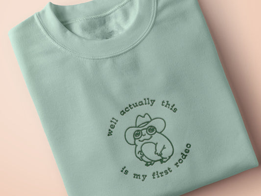An embroidered green t-shirt design of a cute cowboy frog wearing a stetson surrounded by the test well actually this is my first rodeo.
