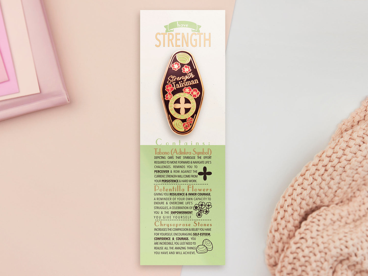 The Strength Talisman gold enamel pin on a gold foiled backing card that outlines the symbolism of the talisman including information and imagery on the Tabono adinkra Symbol, potential flowers, and chrysoprase stones