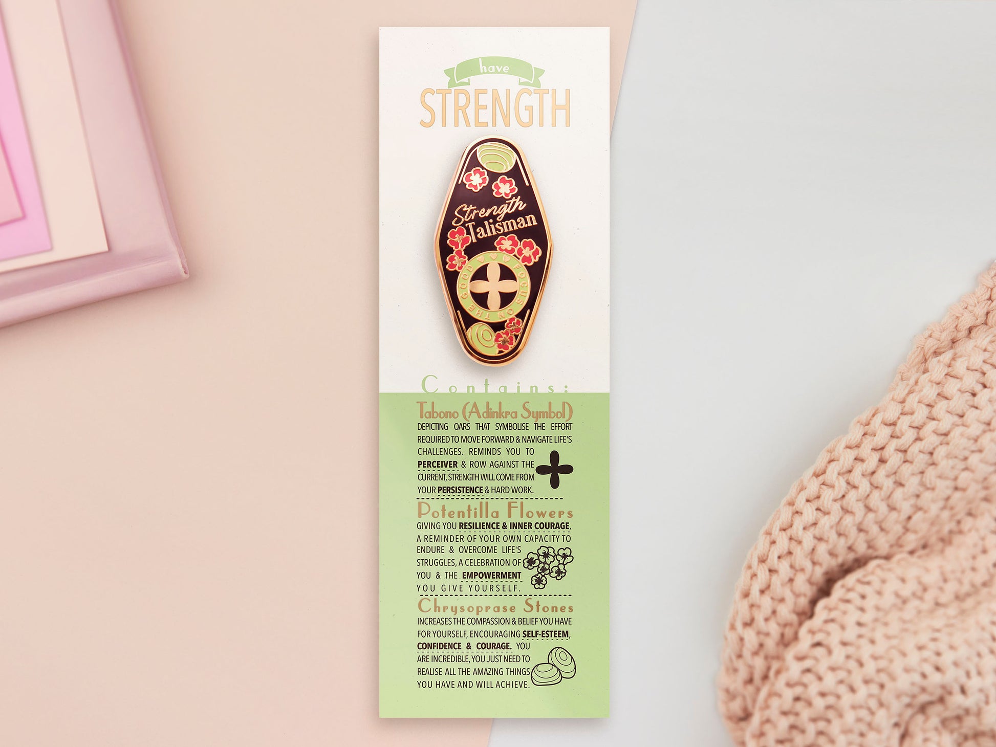 The Strength Talisman gold enamel pin on a gold foiled backing card that outlines the symbolism of the talisman including information and imagery on the Tabono adinkra Symbol, potential flowers, and chrysoprase stones