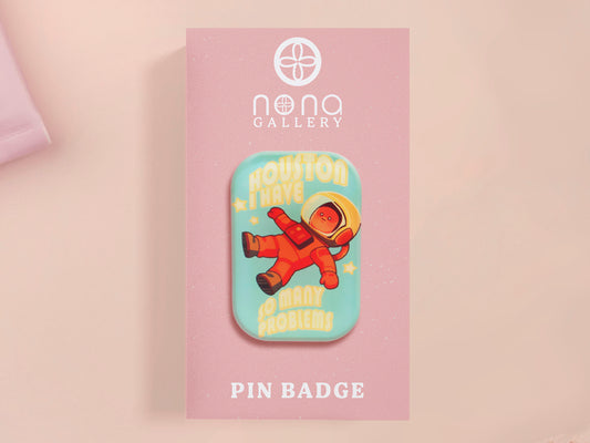 A clear acrylic pin badge with a pink heart-shaped pin back clasp. The design of the pin badge is of a cute chibi retro astronaut floating in sapce with the quote Houston I have so many problems