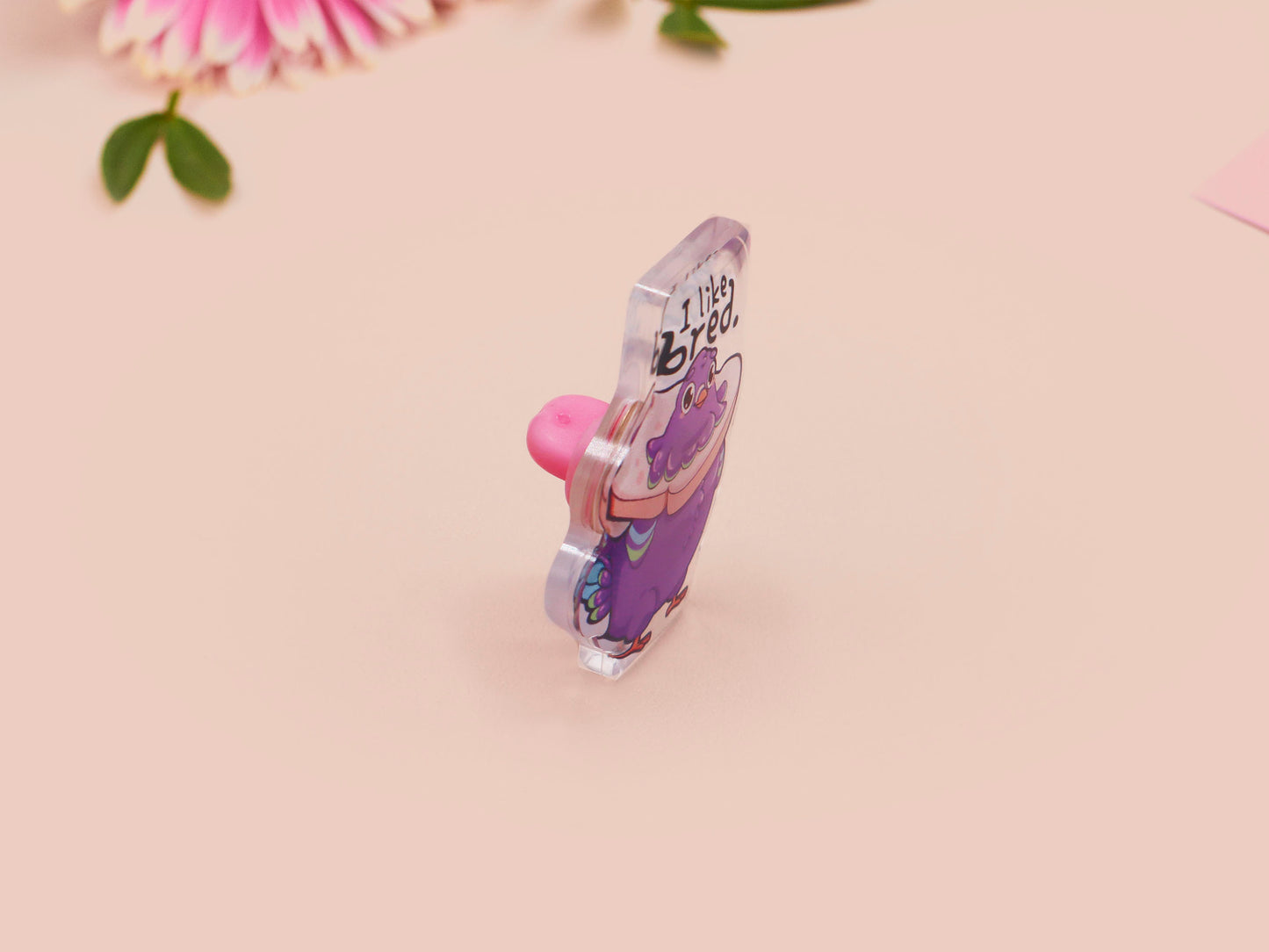 A transparent acrylic pin badge with a pink heart-shaped pin-back clutch. The design of the pin badge is a cute cartoon pigeon with a slice of bread around it's neck with the quote I like bred.