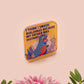 A clear acrylic pin badge with a pink heart-shaped pin back clasp. The design of the pin badge is of a cute chibi blue godzilla like kaiju destroying a city with the quote I Think I Smash Buildings Because My Father Was Distant.