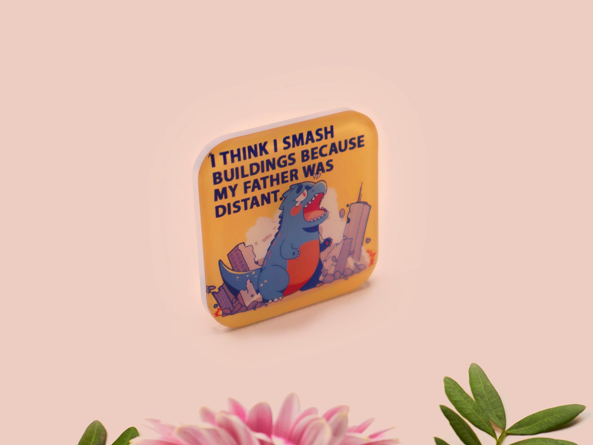 A clear acrylic pin badge with a pink heart-shaped pin back clasp. The design of the pin badge is of a cute chibi blue godzilla like kaiju destroying a city with the quote I Think I Smash Buildings Because My Father Was Distant.