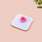 Small pink heart-shaped object on a white square base with a beige background