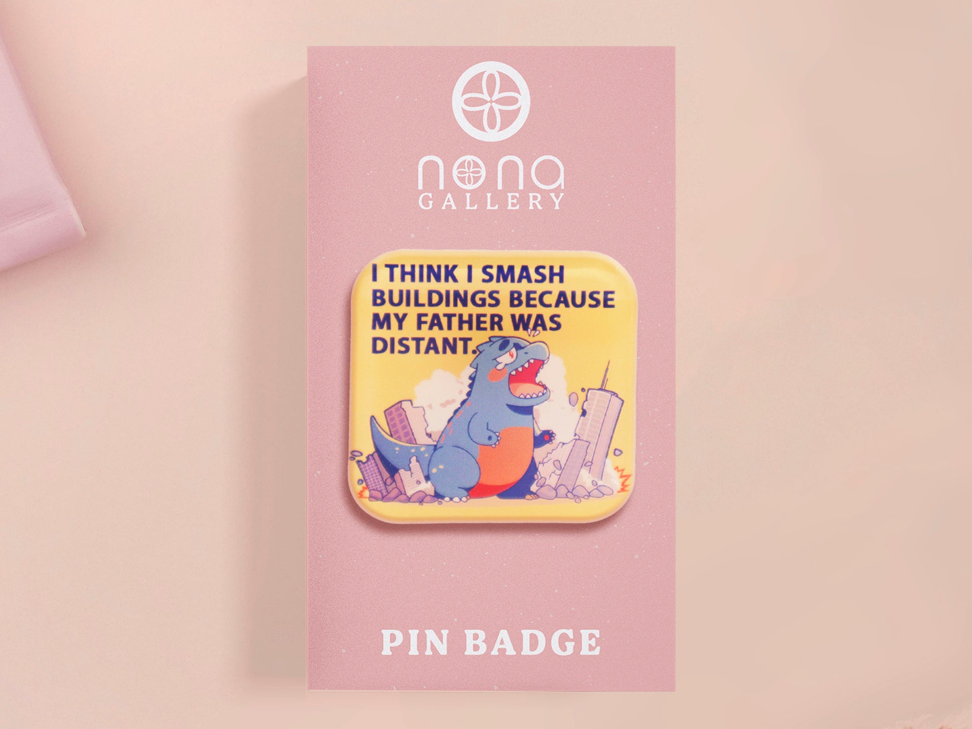 A clear acrylic pin badge with a pink heart-shaped pin back clasp. The design of the pin badge is of a cute chibi blue godzilla like kaiju destroying a city with the quote I Think I Smash Buildings Because My Father Was Distant.