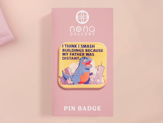 A clear acrylic pin badge with a pink heart-shaped pin back clasp. The design of the pin badge is of a cute chibi blue godzilla like kaiju destroying a city with the quote I Think I Smash Buildings Because My Father Was Distant.