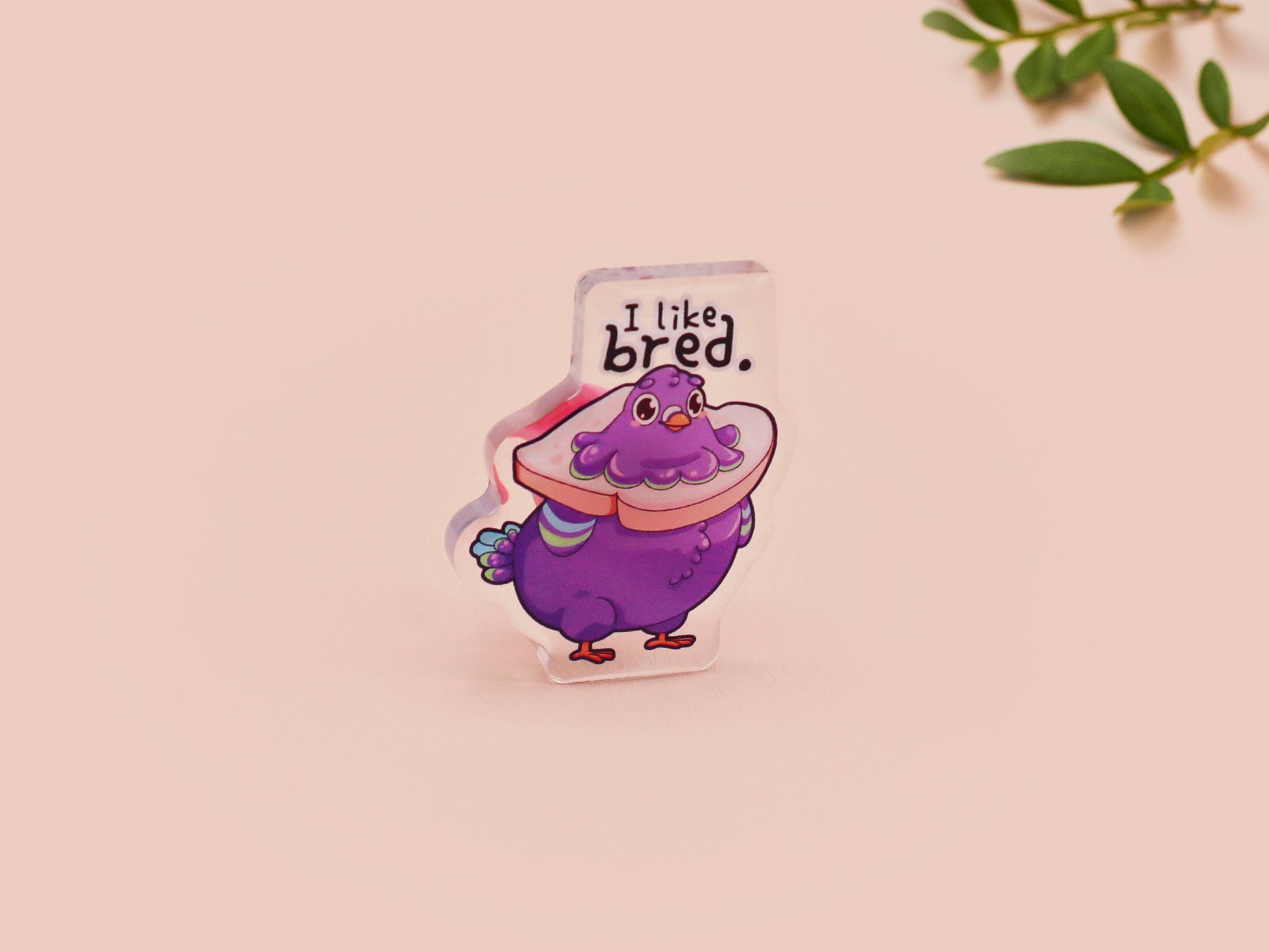 A transparent acrylic pin badge with a pink heart-shaped pin back clutch.The design of the pin badge is a cute cartoon pigeon with a slice of bread around it's neck with the quote I like bred.