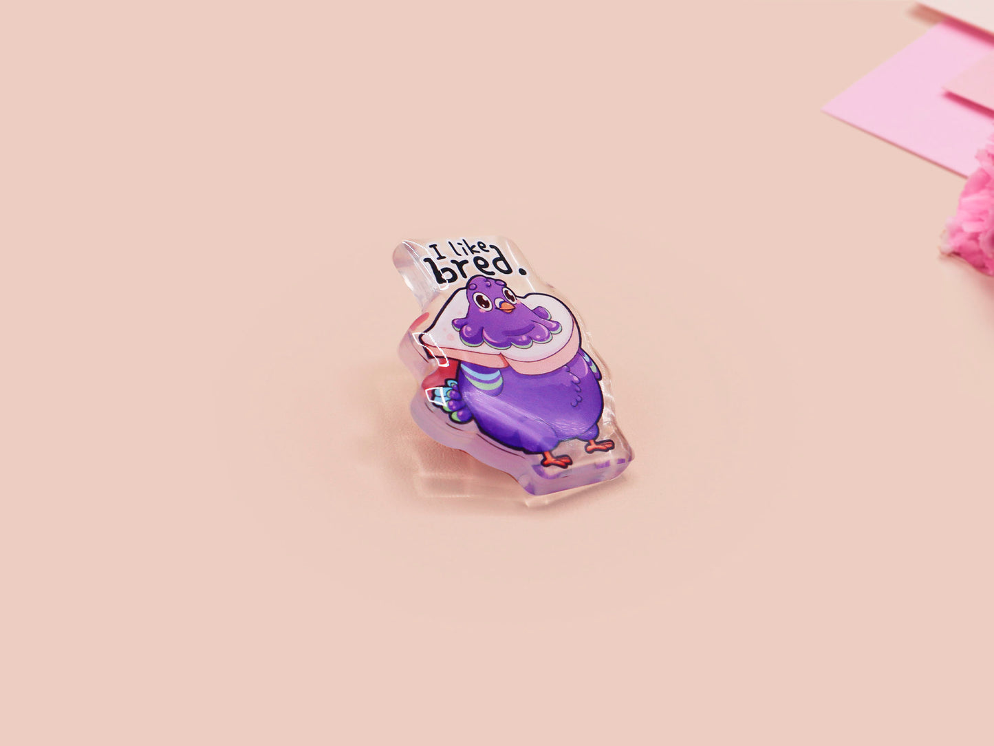 A transparent acrylic pin badge with a pink heart-shaped pin back clutch.The design of the pin badge is a cute cartoon pigeon with a slice of bread around it&#39;s neck with the quote I like bred.