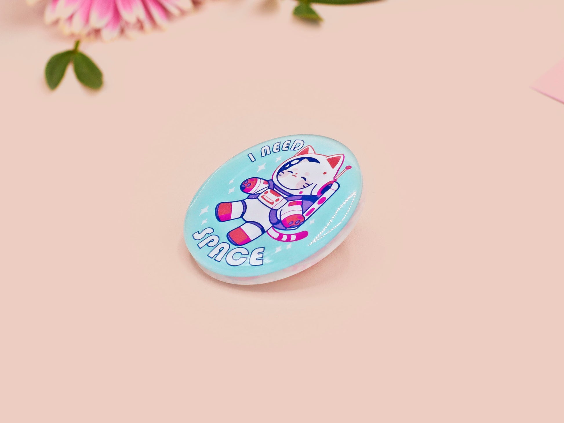 Clear acrylic pin badge with a pink heart-shaped rubber pin back clasp. The design of the pin badge is a cute chibi white cat in a retro astronaut suit with the quote I need space.