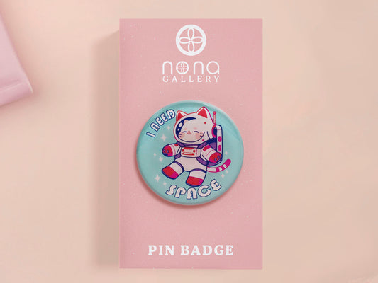 Clear acrylic pin badge with a pink heart-shaped rubber pin back clasp. The design of the pin badge is a cute chibi white cat in a retro astronaut suit with the quote I need space.