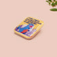 A clear acrylic pin badge with a pink heart-shaped pin back clasp. The design of the pin badge is of a cute chibi blue godzilla like kaiju destroying a city with the quote I Think I Smash Buildings Because My Father Was Distant.