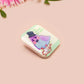 A clear acrylic pin badge with a pink heart-shaped pin back clasp. The design of the pin badge is of a cute chibi pigeon wearing a top hat crying next to a pethetic attempt at nest building with the quote I'm trying my best.
