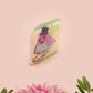 A clear acrylic pin badge with a pink heart-shaped pin back clasp. The design of the pin badge is of a cute chibi pigeon wearing a top hat crying next to a pethetic attempt at nest building with the quote I'm trying my best.