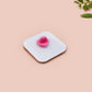 A clear acrylic pin badge with a pink heart-shaped pin back clasp. The design of the pin badge is of a cute chibi pigeon wearing a top hat crying next to a pethetic attempt at nest building with the quote I'm trying my best.