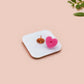 A clear acrylic pin badge with a pink heart-shaped pin back clasp. The design of the pin badge is of a cute chibi pigeon wearing a top hat crying next to a pethetic attempt at nest building with the quote I'm trying my best.