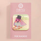 A clear acrylic pin badge with a pink heart-shaped pin back clasp. The design of the pin badge is of a cute chibi pigeon wearing a top hat crying next to a pethetic attempt at nest building with the quote I'm trying my best.