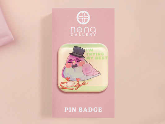 A clear acrylic pin badge with a pink heart-shaped pin back clasp. The design of the pin badge is of a cute chibi pigeon wearing a top hat crying next to a pethetic attempt at nest building with the quote I'm trying my best.