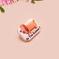 A clear acrylic pin badge with a pink heart-shaped pin back clasp. The design of the pin badge is of a cute chibi dachshund sausage dog with the quote in this economy?