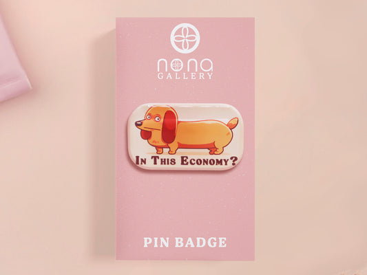 A clear acrylic pin badge with a pink heart-shaped pin back clasp. The design of the pin badge is of a cute chibi dachshund sausage dog with the quote in this economy?
