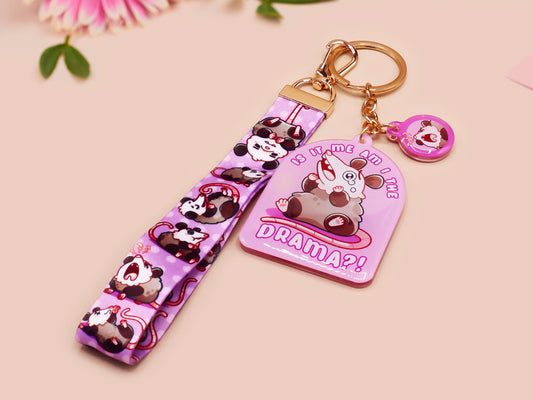 Acrylic epoxy possum keychain with a large acrylic charm of a purple screaming possum with the quote is it me am I the drama, a smaller charm of a cute crying possum and a matching purple possum lanyard.