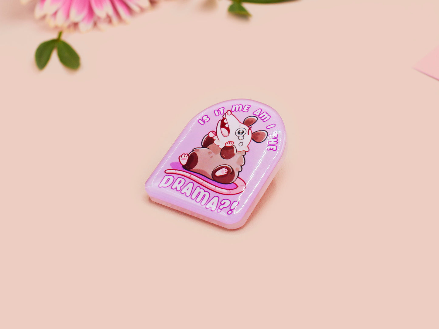 A clear acrylic pin badge with a pink heart-shaped pin back clasp. The design of the pin badge is of a cute screaming possum with the quote is it me am I the drama?