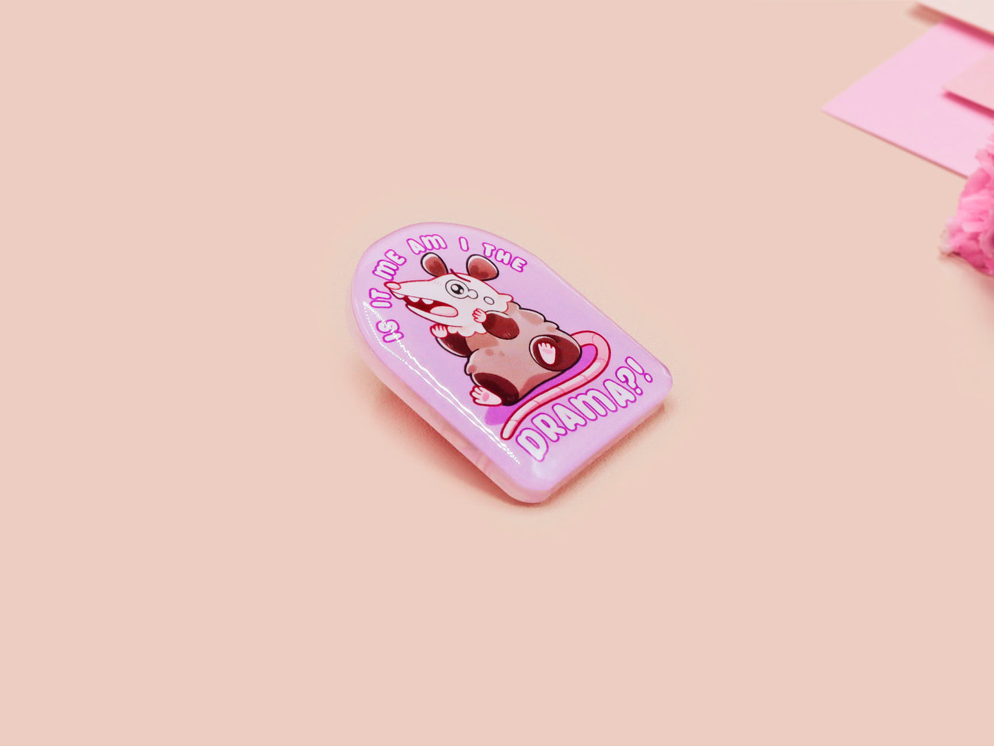 A clear acrylic pin badge with a pink heart-shaped pin back clasp. The design of the pin badge is of a cute screaming possum with the quote is it me am I the drama?