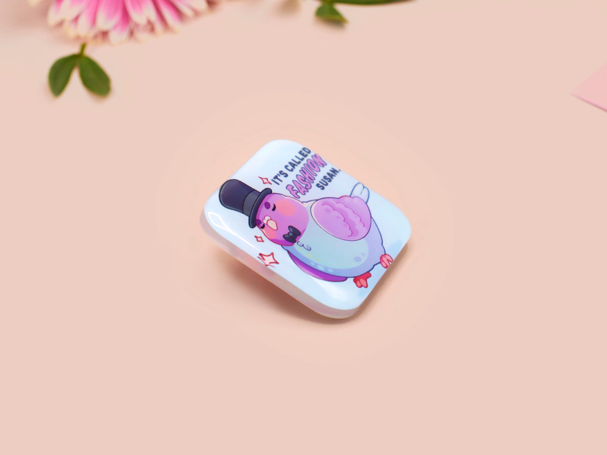 A clear acrylic pin badge with a pink heart-shaped pin back clasp. The design of the pin badge is of a cute chibi pigeon wearing a top hat with the quoteit's called fashion susan