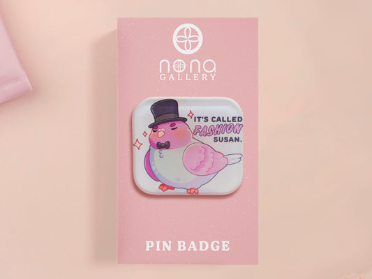 A clear acrylic pin badge with a pink heart-shaped pin back clasp. The design of the pin badge is of a cute chibi pigeon wearing a top hat with the quoteit's called fashion susan